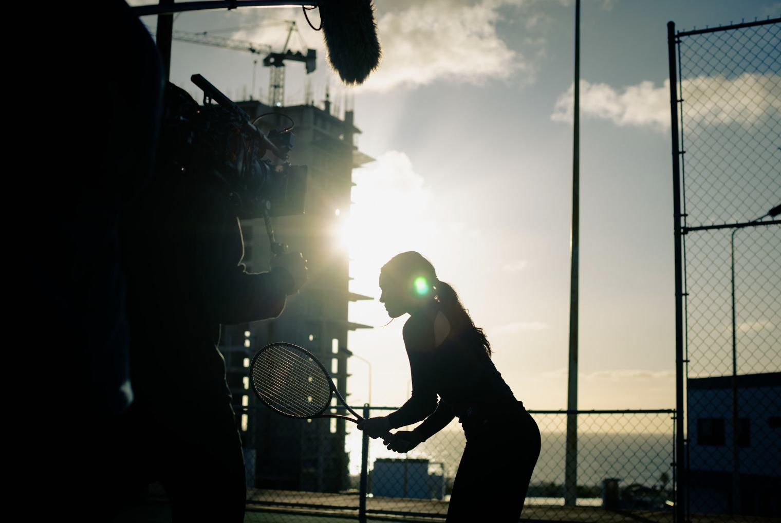 Andrea Londo in silhouette with a tennis racket, backlit by the sun, with a camera operator in the foreground