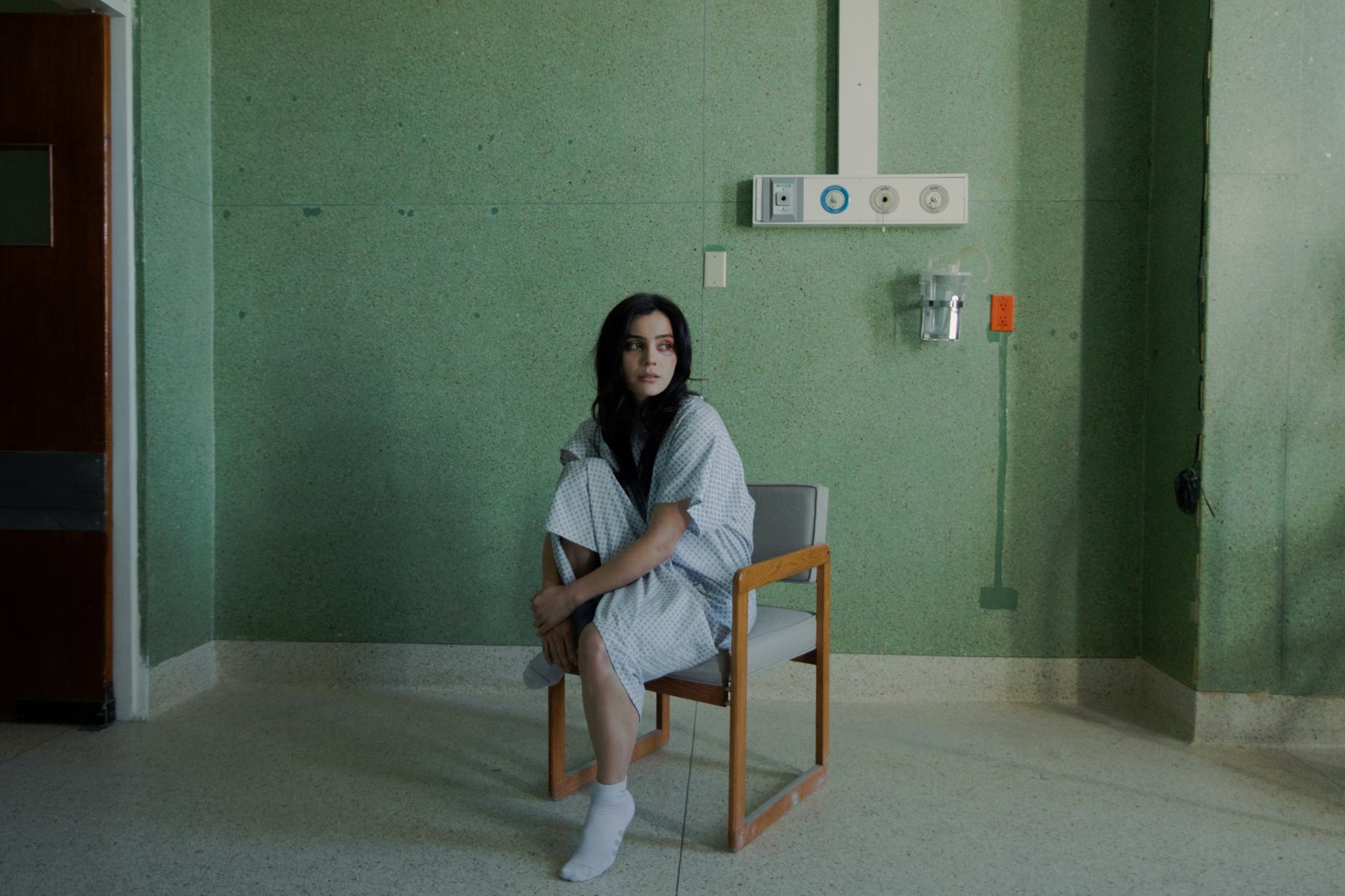 Isa in a hospital gown, sitting alone against a green wall